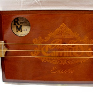 Puede incluir: Una guitarra de caja de puros de madera marrón con letras doradas que dicen "Encore" y "J. Carrillo". La guitarra tiene una etiqueta Reed Made en el lateral.