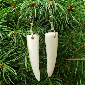 May include: A pair of white bone earrings with silver hooks. The earrings are shaped like long, pointed teeth.