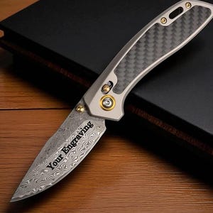 May include: A folding knife with a patterned blade and a gray and black handle. The blade has the text "Your Engraving". The knife is on a wooden surface.