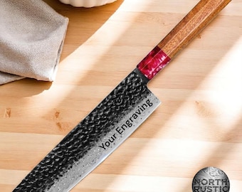 Pode incluir: Uma faca de cozinha artesanal com cabo de madeira e lâmina escura texturizada. O cabo apresenta um detalhe vermelho e a lâmina tem o texto "Your Engraving". A faca é projetada para uso culinário.
