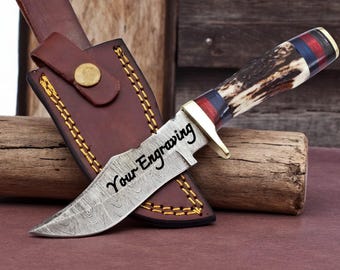 May include: A Damascus steel knife with a brown and white antler handle and a leather sheath. The knife blade has the text "Your Engraving". The sheath is brown leather with gold stitching and a brass button.