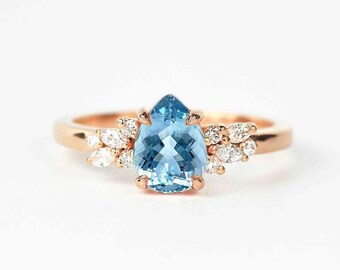 May include: A rose gold ring featuring a pear-shaped, light blue gemstone as the centerpiece. The ring is accented with small, clear gemstones clustered on either side of the main stone, creating a delicate and elegant design. The band is smooth and polished.