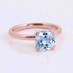 Könnte beinhalten: Roségoldener Ring mit einem quadratischen, hellblauen Edelstein. Der Edelstein wird von vier Krappen gehalten, und der Ring ist aus glattem, poliertem Roségold. Der Ring wird vor weißem Hintergrund gezeigt.