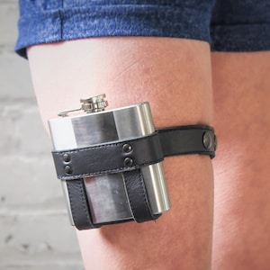 May include: A stainless steel hip flask attached to a thigh with a black leather garter. The flask has a silver cap and is held in place by a black leather strap with rivets. The garter is fastened with a snap.