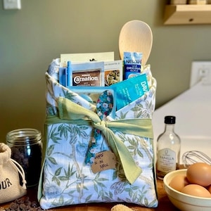 May include: A fabric gift bag with a green and white botanical print, tied with a green ribbon. The bag contains various food items including Carnation and Nescafe packets, a wooden spoon, and a floral patterned spoon with a brown paper tag.
