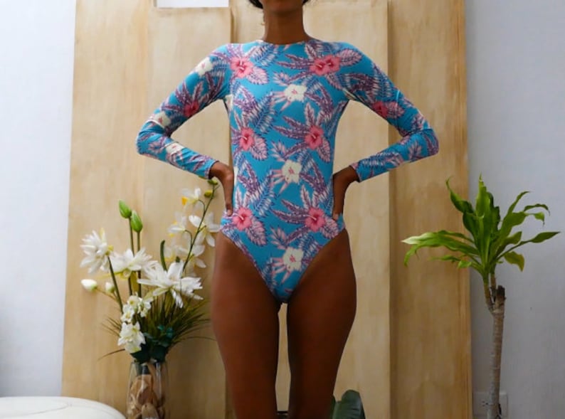 May include: A blue long-sleeved swimsuit with a floral print featuring pink and white flowers. The swimsuit has a scoop neck and a high leg cut.