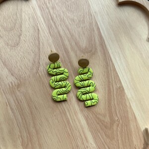 May include: A pair of lime green dangle earrings featuring a wavy, serpentine shape with thin black squiggly line patterns. Each earring is attached to a small, circular, tan-colored stud top, creating a distinct contrast in color and texture.