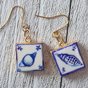 May include: A pair of square earrings featuring a blue and white seashell design. The earrings are made from small, square tiles with gold-coloured hooks. The seashell design is in the centre of the tile, framed by a blue border and floral accents.