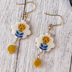Peut inclure: Une paire de boucles d'oreilles dorées. Chaque boucle d'oreille présente un pendentif blanc en forme de fleur avec un motif floral jaune et bleu. Une petite perle jaune pend de chaque pendentif. Les boucles d'oreilles sont posées sur un fond en bois blanc.