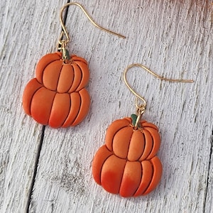 May include: A pair of orange pumpkin earrings with gold-coloured hooks. The earrings are made of a material that resembles clay and have a stacked pumpkin design. The background features dried orange and yellow flowers on a white wooden surface.