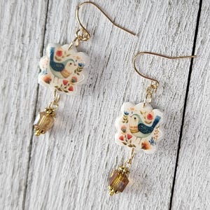 Peut inclure: Une paire de boucles d'oreilles pendantes dorées. Chaque boucle d'oreille présente un charm en forme de fleur blanche avec un oiseau bleu et un motif floral. Une perle facettée de couleur ambre est suspendue en dessous. Les boucles sont sur une surface en bois blanc.