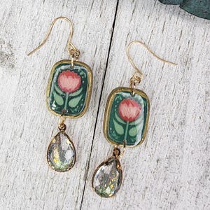 Peut inclure: Une paire de boucles d'oreilles dorées. Chaque boucle d'oreille présente un pendentif rectangulaire avec un motif floral rouge et vert, et un charm en cristal en forme de goutte. Les boucles d'oreilles sont présentées sur une surface en bois blanc.