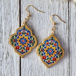 Peut inclure: Boucles d'oreilles pendantes dorées avec un motif floral coloré. Les boucles d'oreilles ont une forme de quadrilobe avec une bordure dorée et présentent un motif de fleurs bleues, jaunes, rouges et vertes.