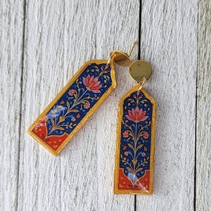 Peut inclure: Une paire de boucles d'oreilles dorées de forme rectangulaire. Les boucles d'oreilles présentent un motif floral détaillé dans des tons de bleu, de rouge et de rose. Le motif est posé sur un fond bleu foncé, avec une bordure dorée. Les boucles d'oreilles sont sur une surface en bois blanc.