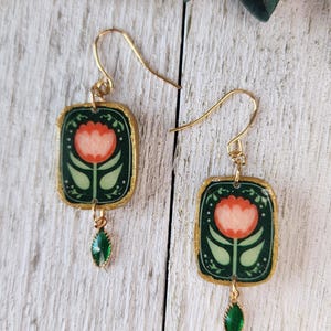 Peut inclure: Une paire de boucles d'oreilles pendantes rectangulaires avec un fond vert foncé et une illustration de tulipe orange au centre. Chaque boucle est encadrée d'or, suspendue à un crochet doré et ornée d'une petite breloque goutte vert émeraude.