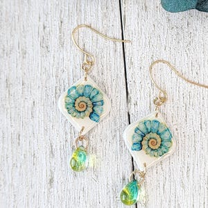 Peut inclure: Une paire de boucles d'oreilles pendantes avec des breloques blanches en forme de losange ornées d'un motif de coquillage en spirale bleu et beige. Chaque boucle comporte un crochet doré et une petite perle verte translucide en forme de goutte.