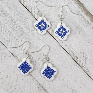Peut inclure: Paire de boucles d'oreilles en céramique bleu et blanc avec un motif géométrique. Les boucles d'oreilles sont sur une surface en bois blanc.