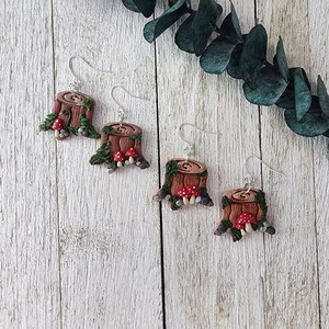 May include: Four pairs of handmade earrings. Each earring features a miniature tree stump design with red mushroom accents, green foliage, and silver-coloured ear wires. The earrings are set against a white wooden background with green leaves.
