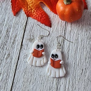 May include: White ghost earrings with black eyes and orange books. The earrings are made of polymer clay and have silver-coloured ear wires. A small orange pumpkin and autumn leaf are in the background.