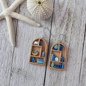 May include: Handmade earrings with a bookshelf design. Each earring depicts a small, arched bookshelf filled with miniature blue books, a vase, and other decorative items. The earrings are set against a weathered, white wooden background.
