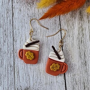 May include: A pair of autumn-themed earrings. Each earring depicts a small orange mug with a white whipped cream topping and a dark brown chocolate stick. A yellow leaf is on the front of the mug. The earrings hang from gold-coloured hooks.