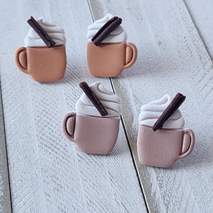 May include: Four miniature coffee mugs with whipped cream and cinnamon sticks. The mugs are in shades of brown and pink, with white whipped cream and dark brown cinnamon sticks. They are arranged on a white wooden surface.