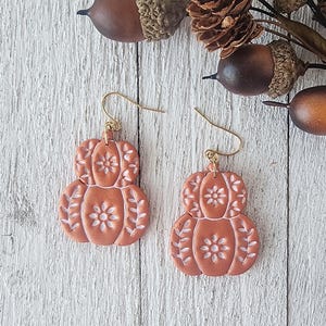May include: A pair of dangle earrings shaped like pumpkins in a matte terracotta orange colour. Each pumpkin features white embossed floral and leaf patterns on the surface and is attached to a gold-toned metal hook for hanging from the ear.