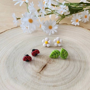 May include: A set of six polymer clay earrings featuring a daisy, a leaf, and a ladybug design. The earrings are displayed on a wooden surface.