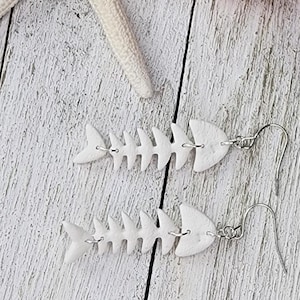 May include: A pair of white fishbone earrings with silver ear wires. The earrings are made of a lightweight material and are designed to resemble a fish skeleton. The earrings are displayed on a white wooden surface.