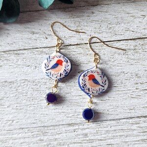 Peut inclure: Une paire de boucles d'oreilles pendantes avec des breloques blanches en forme de losange, ornées d'une illustration peinte d'un oiseau à tête rouge et ailes bleues, entouré de feuilles bleues. Chaque boucle comporte une perle bleu foncé.
