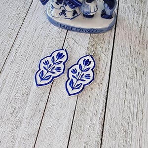 May include: Blue and white Delftware style earrings with a floral design. The earrings are shaped like a leaf and have a silver hook.