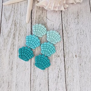 May include: Seashell-shaped earrings in teal and turquoise, with silver hooks. The earrings are displayed on a light wooden surface, with a starfish and seashell in the background. The earrings are a beach-themed accessory.