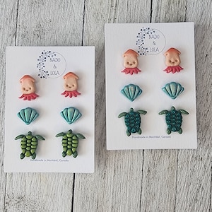 May include: Two sets of sea-themed earrings displayed on white cards. Each set features a pair of octopus earrings, a pair of seashell earrings, and a pair of sea turtle earrings. The octopus earrings are peach with red tentacles. The seashells are light blue with a green iridescent sheen. The sea turtles are green with a darker green shell. The cards include the text "NADO & LOLA" and "Handmade in Montreal, Canada".
