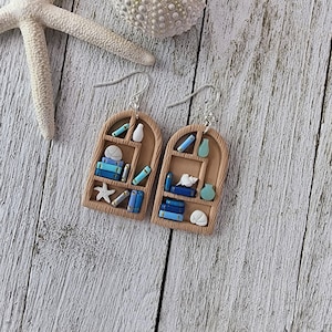 May include: Handmade earrings in the shape of a miniature bookshelf. The brown shelves are filled with tiny books, vases, and a starfish. The earrings hang from silver hooks. A starfish and sea urchin are also visible in the image.