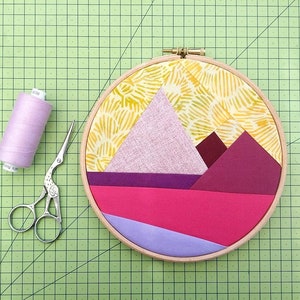Pode incluir: Uma cena de montanha de tecido roxo, rosa e vermelho em um aro de bordado de madeira. O aro está em um tapete de corte verde com um carretel de fio lavanda e um par de tesouras prateadas.