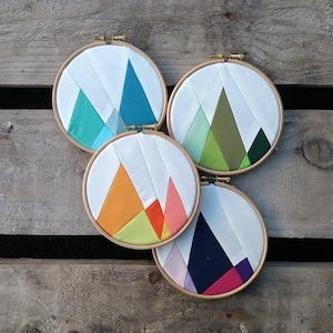 May include: Four circular embroidery hoops with colorful geometric mountain designs. The hoops are made of wood and have a white background. The mountains are made of fabric in various colors, including blue, green, orange, and pink.