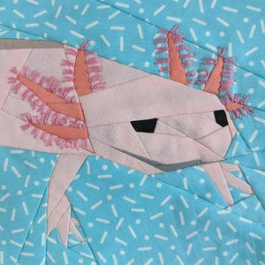 Op de afbeelding: Een roze en witte stoffen axolotl met roze gevederde kieuwen, gestikt in een patchwork-stijl. De axolotl bevindt zich op een blauwe achtergrond met witte streepjes.