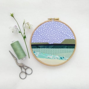 Może przedstawiać: Ręcznie wykonane dzieło sztuki tekstylnej w drewnianym tamborku. Grafika przedstawia warstwy tkaniny w odcieniach niebieskiego, zielonego i kremowego, tworząc krajobraz. Szpula zielonej nici i małe nożyczki są umieszczone w pobliżu.