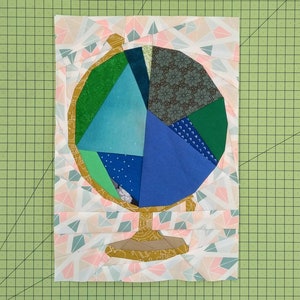 Peut inclure: Un globe en patchwork avec un fond blanc et un pied doré. Le globe est fait de tissus de différentes couleurs, notamment du vert, du bleu et du marron. Le globe est entouré d'un fond blanc avec des formes géométriques roses et bleues.