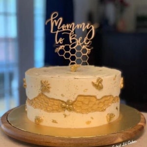 May include: A white cake decorated with gold honeycomb and bee accents. The cake is topped with a wooden cake topper that says "Mommy to Bee".