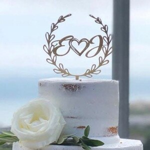 May include: A three-tiered white wedding cake decorated with olive branches and a wooden topper with the initials "E & J" in a wreath. The cake is on a dark brown cake stand.