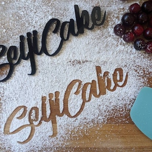 May include: Two black acrylic "Seiji Cake" cutouts, one solid and one negative space, on a wooden surface dusted with flour. Fresh cherries and a teal spatula are also present, suggesting a baking theme.