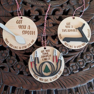 May include: Three wooden ornaments with red and white string. One ornament has a spoon, another a treadmill, and the third has jumper cables. Each ornament has a phrase: "Got you a spoon, since you like to stir the pot", "Got you a treadmill, you like to run you", and "Got you some jumper cables, since you like to start shit".