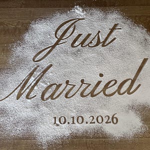 May include: A wooden surface with the words "Just Married" and the date "10.10.2026" written in white powder. The text is in a cursive font, creating a romantic and celebratory feel. Ideal for wedding announcements.