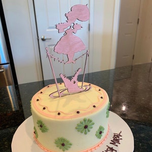 May include: A white cake with pink and green frosting decorations. A wooden cutout of a girl jumping rope over an alligator is on top of the cake. The cake is on a white plate with the words "Happy" written on it.