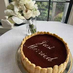 May include: A round tiramisu cake surrounded by ladyfinger cookies, topped with cocoa powder and the text "Just Married" in white script. A clear glass vase filled with white orchids sits behind the cake on a white textured tablecloth.
