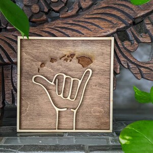 May include: A wooden sign with a brown background and a light brown outline. The sign features a hand giving the "hang loose" gesture with the outline of the Hawaiian islands inside the hand.