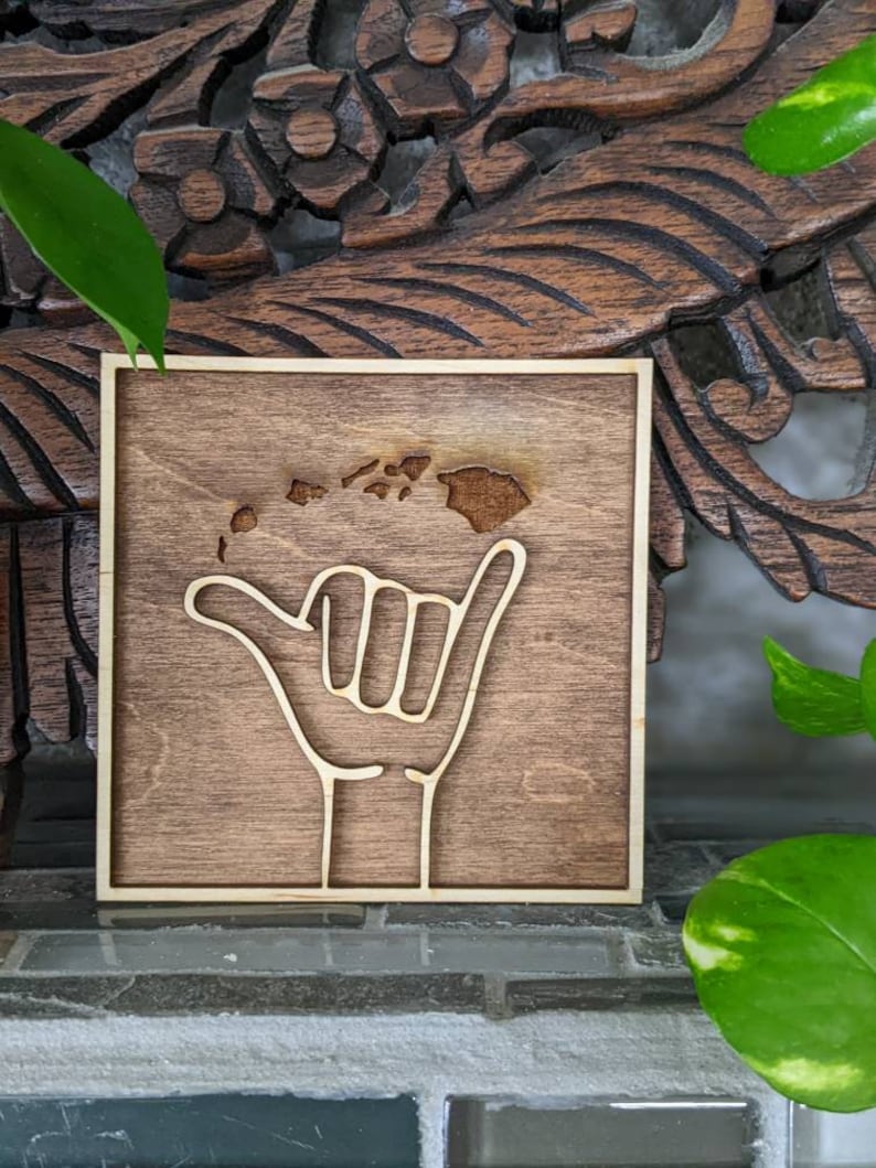 May include: A wooden sign with a brown background and a light brown outline. The sign features a hand giving the "hang loose" gesture with the outline of the Hawaiian islands inside the hand.