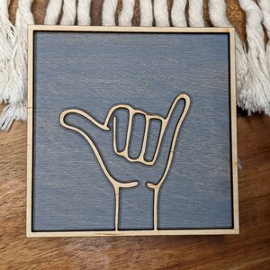 May include: A gray wooden sign with a white outline of a hand giving the shaka sign. The sign is a square shape and has a light wood frame.