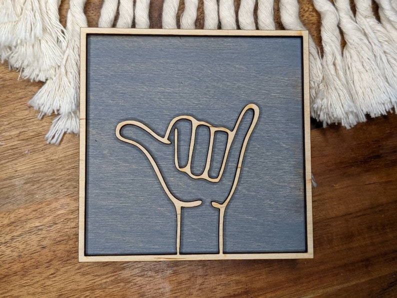 May include: A gray wooden sign with a white outline of a hand giving the shaka sign. The sign is a square shape and has a light wood frame.